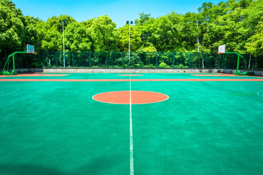 Empty outdoor basketball court with painted lines and a center circle, bordered by a chain-link fence and trees