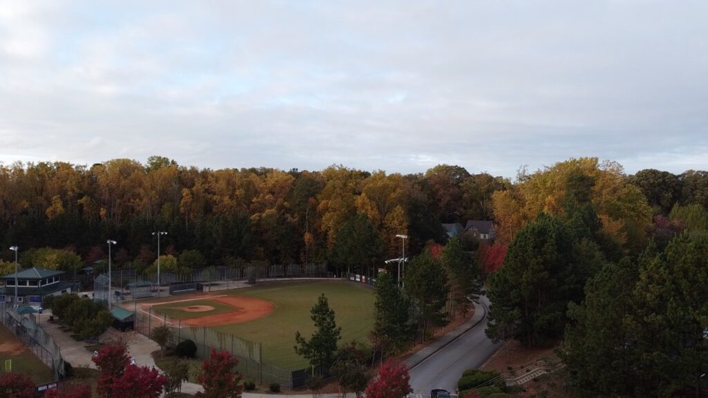 Aerial view of an empty baseball diamond with a dirt infield and grass outfield, framed by autumn trees