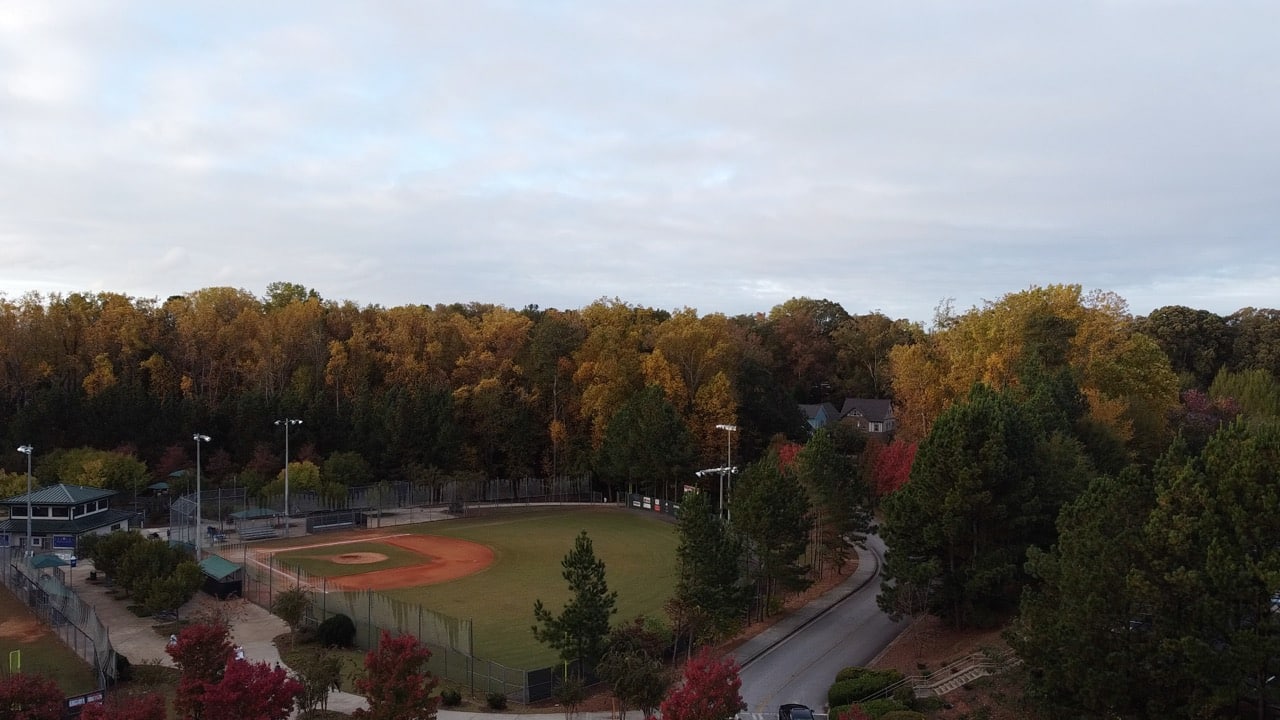 Aerial view of an empty baseball diamond with a dirt infield and grass outfield, framed by autumn trees